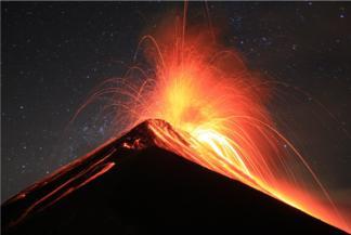 Erupting volcano in front of a night sky with stars. Fountains of white-yellow-orangered
magma erupt from the summit of the volcano, and the lava can be seen to be running down
the flanks of the volcano, which is mostly black as the photograph is taken at night.