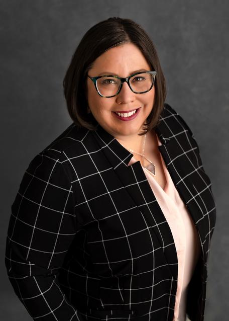 A light-skinned woman with brown hair and glasses wearing a black suit jacket with a white square pattern and a light pink shirt.